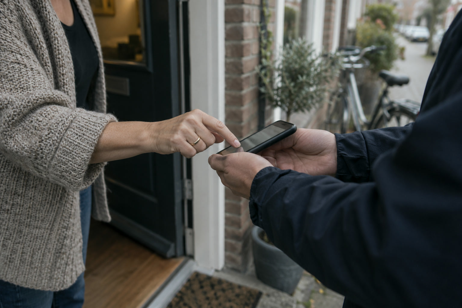 Een vrouw in een gebreid vest tekent bij haar voordeur met haar vinger op de telefoon van een monteur in donkerblauwe werkkleding.
